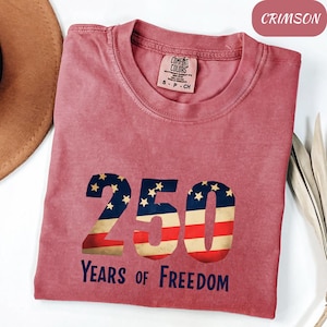 May include: Crimson t-shirt with a patriotic "250" design, incorporating stars and stripes, and the words "Years of Freedom" in navy blue. The shirt has a crew neck and a small tag near the collar. The shirt is a size S-P-CH.