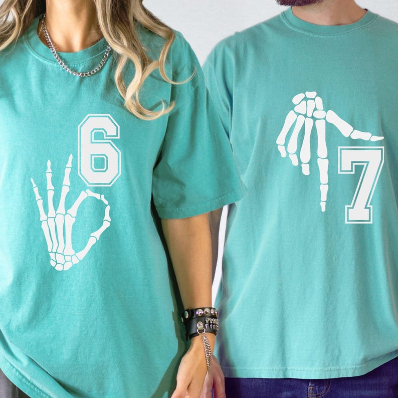 May include: Teal-colored t-shirts with white skeleton hand graphics. One shirt displays the number 6 with an OK hand gesture, and the other shows the number 7 with a hand gesture. The shirts are short-sleeved.