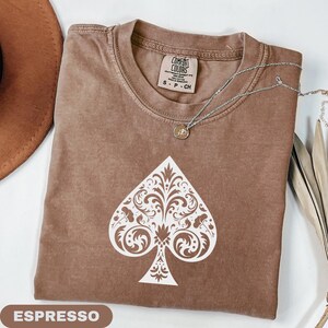 Puede incluir: Camiseta color espresso con un dise&ntilde;o de pica blanco ornamentado. La camiseta tiene cuello redondo y mangas cortas. Un collar plateado con un peque&ntilde;o colgante est&aacute; drapeado sobre la camiseta. La palabra "ESPRESSO" se muestra en un rect&aacute;ngulo marr&oacute;n en la parte inferior.