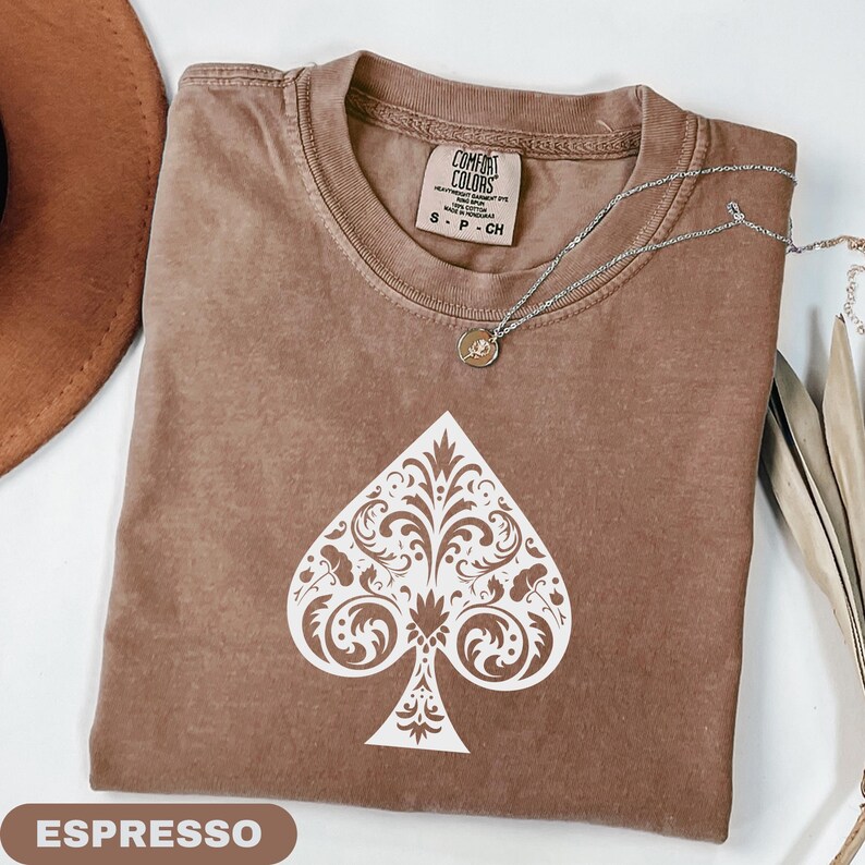Puede incluir: Camiseta color espresso con un dise&ntilde;o de pica blanco ornamentado. La camiseta tiene cuello redondo y mangas cortas. Un collar plateado con un peque&ntilde;o colgante est&aacute; drapeado sobre la camiseta. La palabra "ESPRESSO" se muestra en un rect&aacute;ngulo marr&oacute;n en la parte inferior.