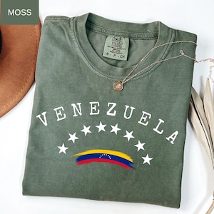 Op de afbeelding: Een mosgroen t-shirt met het woord "VENEZUELA" in witte letters, gebogen boven witte sterren en een gestileerde Venezolaanse vlag. Een gouden ketting met een hanger ligt op het shirt. Comfort Colors merk.