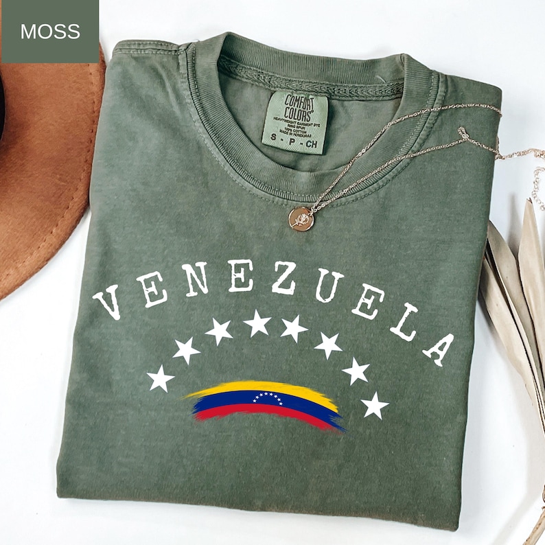 Op de afbeelding: Een mosgroen t-shirt met het woord "VENEZUELA" in witte letters, gebogen boven witte sterren en een gestileerde Venezolaanse vlag. Een gouden ketting met een hanger ligt op het shirt. Comfort Colors merk.