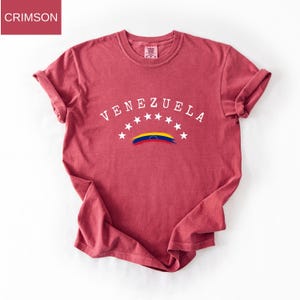 Op de afbeelding: Karmizijnrood T-shirt met het woord "VENEZUELA" in wit, gebogen over de borst. Daaronder staan zeven witte sterren en een gestileerde Venezolaanse vlag. Het shirt heeft opgerolde mouwen.