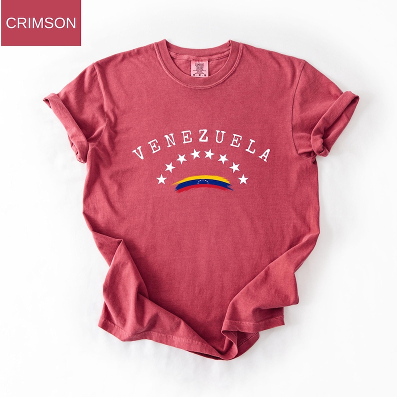 Op de afbeelding: Karmizijnrood T-shirt met het woord "VENEZUELA" in wit, gebogen over de borst. Daaronder staan zeven witte sterren en een gestileerde Venezolaanse vlag. Het shirt heeft opgerolde mouwen.