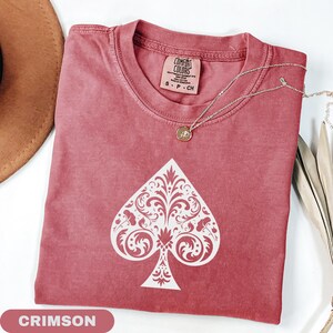Puede incluir: Camiseta de color carmes&iacute; con un dise&ntilde;o de pica blanco. La camiseta tiene cuello redondo y mangas cortas. Un collar plateado con un peque&ntilde;o colgante est&aacute; drapeado sobre la camiseta. La camiseta est&aacute; etiquetada como "Comfort Colors" y es talla S.