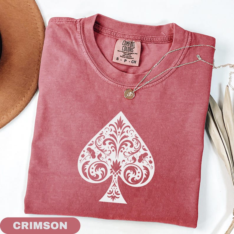 Puede incluir: Camiseta de color carmes&iacute; con un dise&ntilde;o de pica blanco. La camiseta tiene cuello redondo y mangas cortas. Un collar plateado con un peque&ntilde;o colgante est&aacute; drapeado sobre la camiseta. La camiseta est&aacute; etiquetada como "Comfort Colors" y es talla S.