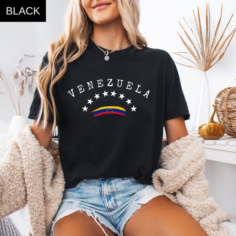 Op de afbeelding: Zwart T-shirt met het woord "VENEZUELA" boven een ontwerp van de Venezolaanse vlag en sterren. Het T-shirt heeft een ronde hals en wordt gedragen met een spijkerbroek en een cr&egrave;mekleurig vest.