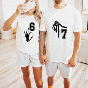 May include: Two white t-shirts with black skeleton hand graphics and numbers 6 and 7. The shirts are paired with gray shorts. The image shows a couple holding hands, with one person holding a phone.