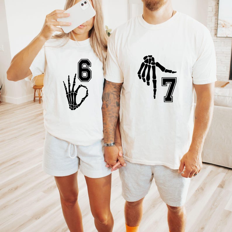 May include: Two white t-shirts with black skeleton hand graphics and numbers 6 and 7. The shirts are paired with gray shorts. The image shows a couple holding hands, with one person holding a phone.