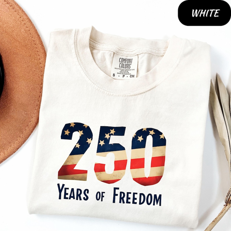 May include: Cream-coloured t-shirt with the number "250" in a patriotic design, featuring red and white stripes and blue stars. The text "YEARS OF FREEDOM" is below the number. The shirt is folded.