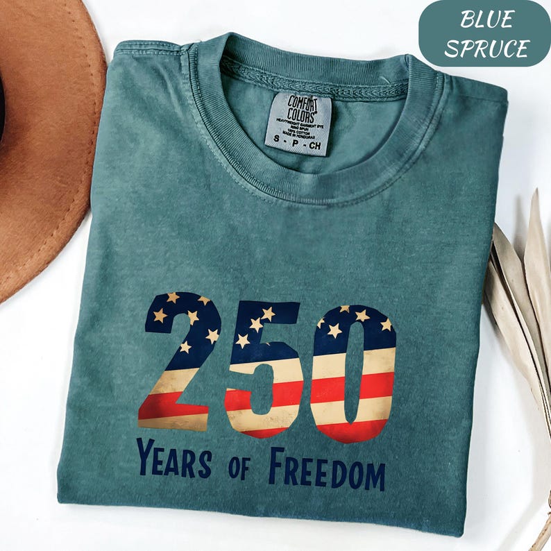 May include: Blue spruce t-shirt with the number 250 in a patriotic design, featuring stars and stripes. The text "Years of Freedom" is printed below the number. The shirt has a Comfort Colors tag.
