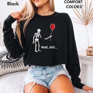 May include: A black long-sleeve sweatshirt featuring a white graphic of a skeleton holding a red balloon with one detached arm. The text "Well, shit..." is printed in white below the skeleton. The garment is labeled as Comfort Colors brand.