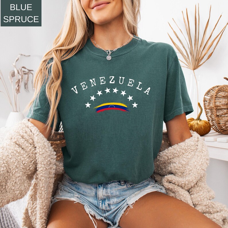 Op de afbeelding: Blauwspar T-shirt met het woord "VENEZUELA" in witte gebogen tekst boven een Venezolaanse vlagafbeelding. Het shirt heeft een ronde hals en korte mouwen. De tekst "BLUE SPRUCE" staat in een vakje linksboven.
