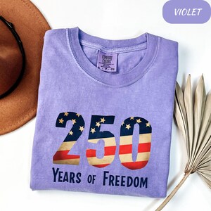 May include: A violet Comfort Colors t-shirt with the number "250" in a patriotic design, featuring stars and stripes. The text "Years of Freedom" is printed below. The shirt has a crew neck and a small tag that says "Comfort Colors".