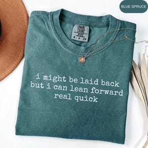 Puede incluir: Camiseta de color azul abeto con el texto "i might be laid back but i can lean forward real quick" en blanco. Un collar dorado con un colgante está drapeado sobre la camiseta. La camiseta está doblada sobre una superficie blanca.