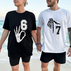 May include: Black and white t-shirts with skeleton hand graphics. One shirt displays the number 6 with an OK hand gesture, the other shows the number 7 with a similar hand design. Both shirts are short-sleeved.