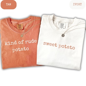 May include: Two t-shirts, one yam-colored and one ivory, with the phrases "kind of rude potato" and "sweet potato" printed on them. Each shirt has a Comfort Colors tag and a gold necklace with a pendant.