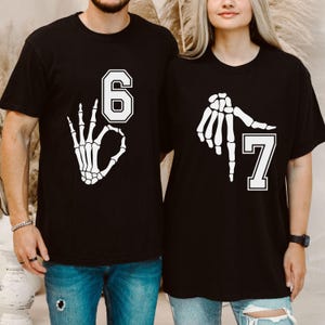 May include: Black t-shirts with white skeleton hand graphics. One shirt displays the number 6 with an 'okay' hand gesture, and the other shows the number 7 with a pointing hand. The shirts are likely for a couple or group.