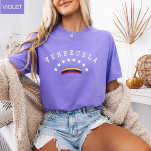 Op de afbeelding: Een violetkleurig T-shirt met het woord "VENEZUELA" in witte letters over de borst, boven een gestileerde Venezolaanse vlag. Het shirt heeft korte mouwen en is gemaakt van een zacht materiaal. Het woord "VIOLET" staat in een vakje linksboven.