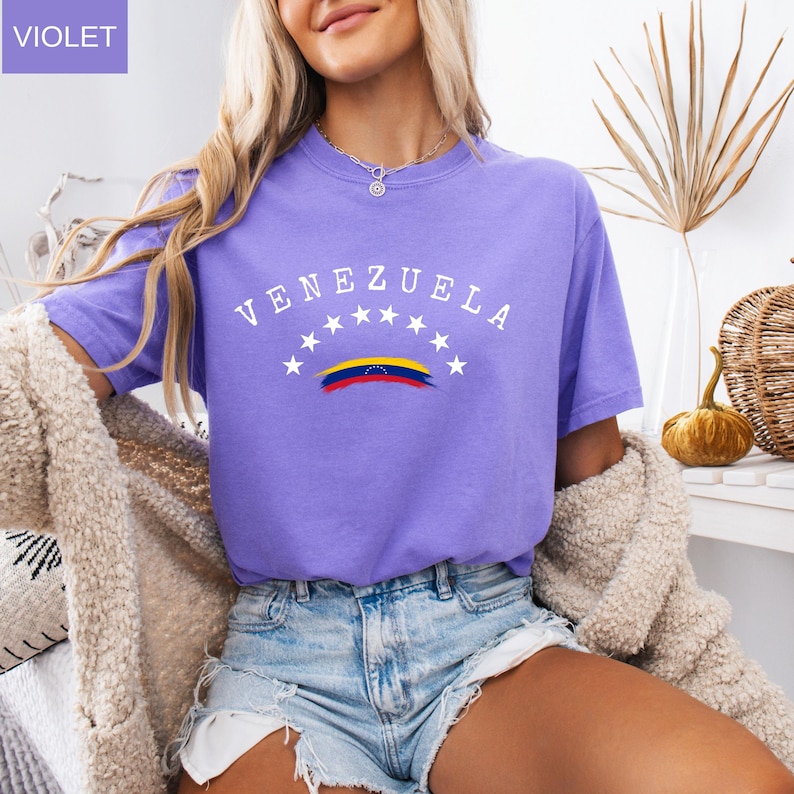 Op de afbeelding: Een violetkleurig T-shirt met het woord "VENEZUELA" in witte letters over de borst, boven een gestileerde Venezolaanse vlag. Het shirt heeft korte mouwen en is gemaakt van een zacht materiaal. Het woord "VIOLET" staat in een vakje linksboven.