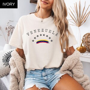 Op de afbeelding: Ivoor T-shirt met het woord "VENEZUELA" in een boog, boven een rij sterren en een Venezolaanse vlagafbeelding. Het shirt met korte mouwen heeft een ronde hals. Het woord "IVORY" staat in een zwart kader linksboven.