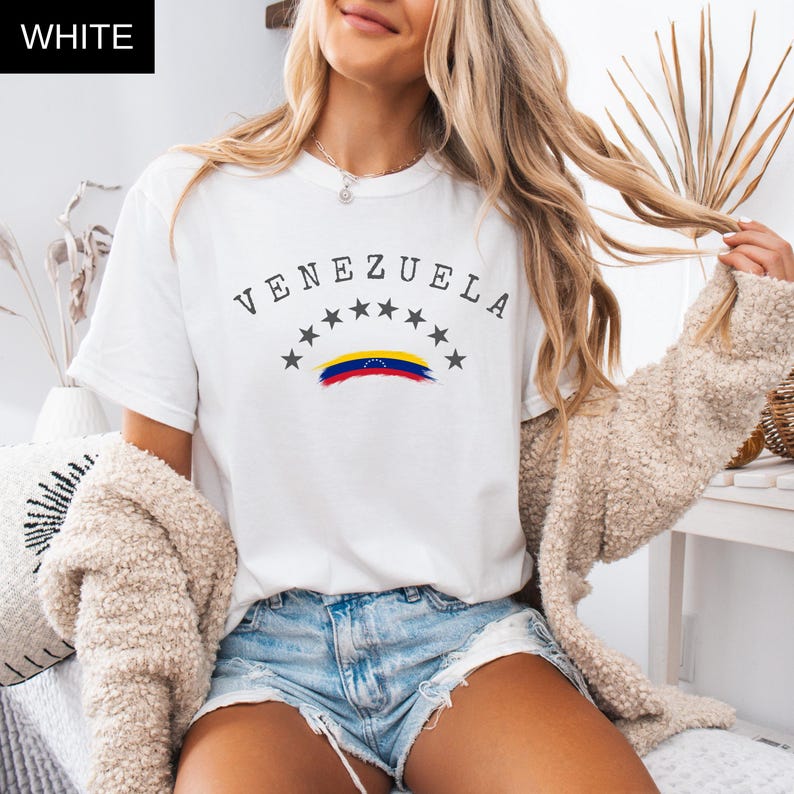 Op de afbeelding: Wit T-shirt met het woord "VENEZUELA" in een boog, boven sterren en een gestileerde Venezolaanse vlag. Het T-shirt heeft een ronde hals.