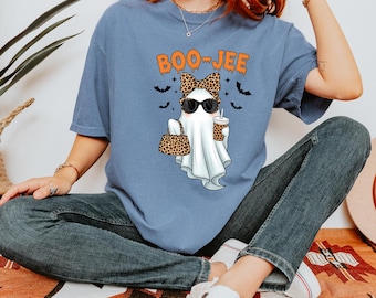 以下が含まれることがあります： オレンジ色の「BOO-JEE」の文字が入った水色のTシャツ。グラフィックは、サングラスをかけ、ヒョウ柄のリボンをつけ、ヒョウ柄のハンドバッグとコーヒーカップを持ったお化けが特徴です。黒いコウモリもデザインに含まれています。
