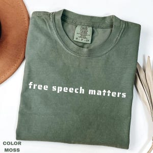 May include: A moss green t-shirt with the white text "free speech matters". The shirt is folded and features a Comfort Colors label. A brown hat and dried leaves are partially visible in the background.
