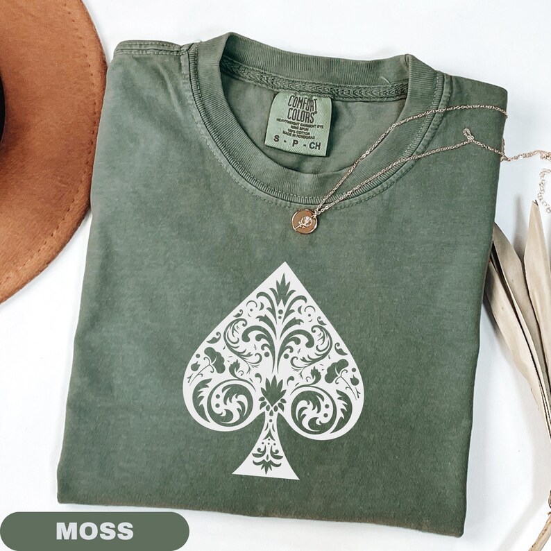 Puede incluir: Una camiseta verde musgo con un dise&ntilde;o de pica blanco. La pica est&aacute; llena de un patr&oacute;n floral adornado. Un collar dorado con un peque&ntilde;o colgante descansa sobre la camisa. La camisa est&aacute; doblada y exhibida sobre una superficie blanca.