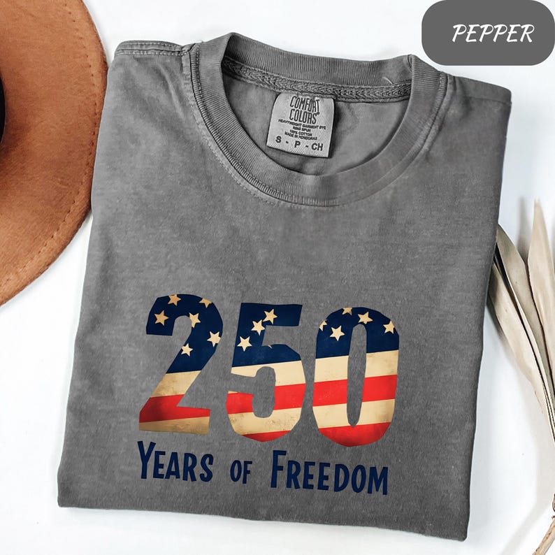 May include: A grey Comfort Colors t-shirt featuring the number 250 in a patriotic design with red, white, and blue stripes and stars. The text "Years of Freedom" is below. The shirt is folded.