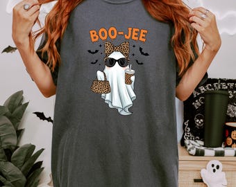 以下が含まれることがあります： ハロウィーンのゴーストグラフィックが描かれた濃いグレーのTシャツ。 ゴーストはサングラス、ヒョウ柄のリボンを着用し、お揃いのハンドバッグとコーヒーカップを持っています。 「BOO-JEE」の文字がオレンジ色でゴーストの上にあります。