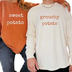May include: Two long-sleeved shirts, one orange and one cream-colored, are displayed. The orange shirt has the words "sweet potato" in white, while the cream shirt says "grouchy potato" in orange. Both shirts are casual wear.