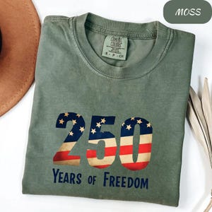 May include: A moss green t-shirt featuring the number "250" in a design inspired by the American flag. The text "Years of Freedom" is below. The shirt is made of a soft material.