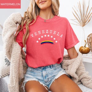 Op de afbeelding: Een watermeloenkleurig T-shirt met het woord "VENEZUELA" in het wit, gebogen boven een rij witte sterren en een Venezolaanse vlagafbeelding. Het shirt heeft korte mouwen en een ronde hals.