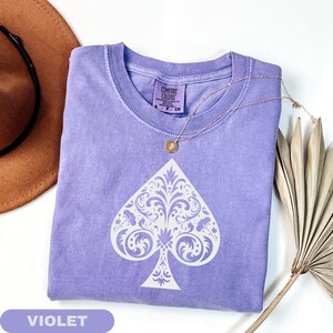 Puede incluir: Camiseta violeta con un dise&ntilde;o de pica blanco. La pica est&aacute; intrincadamente estampada con detalles florales y de volutas. La camiseta tiene cuello redondo y mangas cortas. Un collar dorado con un colgante descansa sobre la camiseta. La etiqueta dice "Comfort Colors".