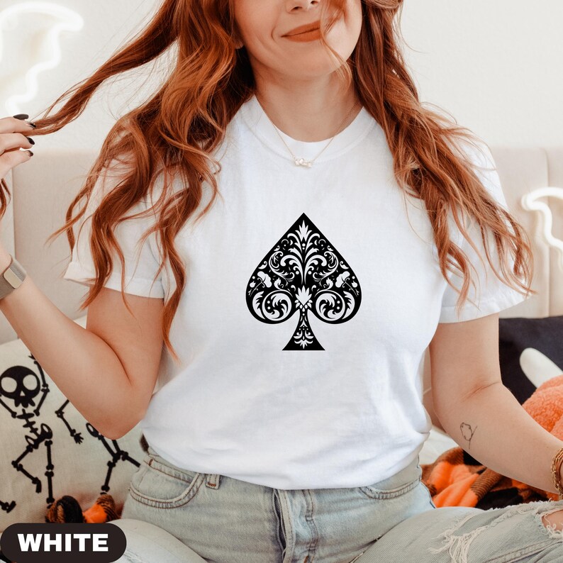 Puede incluir: Camiseta blanca con un dise&ntilde;o de pica negra adornada. La pica est&aacute; llena de patrones florales arremolinados. La palabra "WHITE" es visible en la parte inferior de la imagen. Una prenda casual para el d&iacute;a a d&iacute;a.