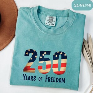 May include: Seafoam green t-shirt with a patriotic "250" design, incorporating stars and stripes. The text "Years of Freedom" is below. The shirt has a crew neck and short sleeves. The label reads "Comfort Colors".