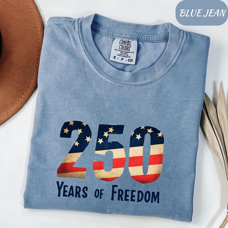 May include: A pale blue t-shirt featuring the text "250 Years of Freedom". The numbers are designed with the American flag, incorporating stars and stripes. The shirt is a Comfort Colors brand.