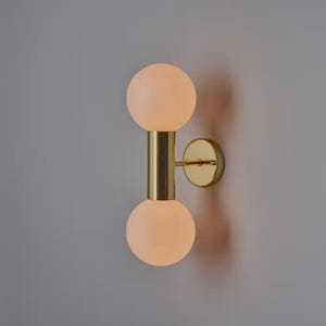 Puede incluir: Aplique de pared moderno con acabado en latón y dos pantallas esféricas de vidrio esmerilado. La luminaria está montada en una pared, mostrando un diseño minimalista. El aplique es una elegante lámpara.