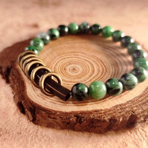 Puede incluir: Una pulsera de cuentas redondas verdes y negras. Cuenta con anillos de metal dorados y un cierre marrón oscuro. La joya se exhibe sobre una rodaja de madera, mostrando la veta natural.