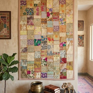 May include: A colourful patchwork wall hanging made of various patterned fabric squares in yellow, orange, green, and purple. The hanging has a pink border and is mounted on a beige wall. A dark wooden table holds a brass pot, books, and an elephant figurine.