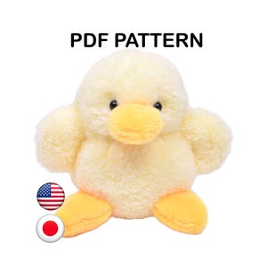 Può includere: Un peluche a forma di anatroccolo giallo con una consistenza morbida e soffice, occhi scuri e becco e zampe di colore arancione brillante. Il testo PDF PATTERN è visualizzato sopra l'anatroccolo, con piccole icone circolari delle bandiere americana e giapponese.