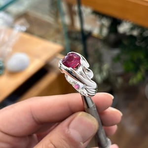 May include: A silver ring featuring a large, oval-cut ruby gemstone. The ring has a detailed, flowing design around the ruby, with smaller accent stones. The ring is held by a tool, showcasing the craftsmanship and design.