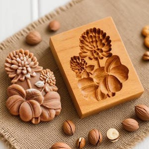 May include: A wooden cookie mold with a floral design and a baked cookie. The mold is rectangular and features a carved flower and leaf pattern. The cookies are round and have a similar floral design. Walnuts are scattered around the mold.