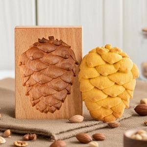 May include: A wooden pine cone cookie mold next to a baked pine cone-shaped cookie. The mold is made of light brown wood with a carved pine cone design. The cookie is golden yellow and has a detailed pine cone shape. Nuts are scattered around.