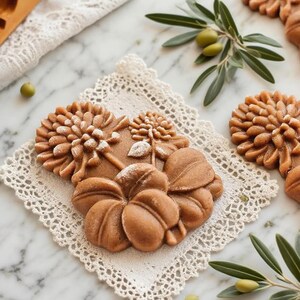 May include: Close-up of intricately designed brown cookies, shaped like flowers and leaves, resting on a white lace doily. The cookies are dusted with a light powder, and olive branches with green olives are scattered around.