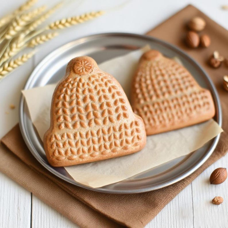 May include: Two baked goods shaped like winter hats sit on a silver plate with parchment paper. The hats are light brown with detailed leaf-like patterns. Wheat stalks and nuts are scattered around the plate on a brown cloth, suggesting a bakery or food theme.