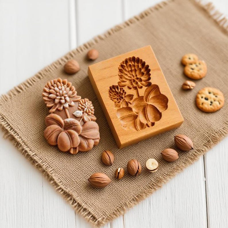 May include: A wooden cookie mold with a floral design and a baked cookie. The mold is rectangular and features a carved flower and leaf pattern. The cookies are round and have a similar floral design. Walnuts are scattered around the mold.
