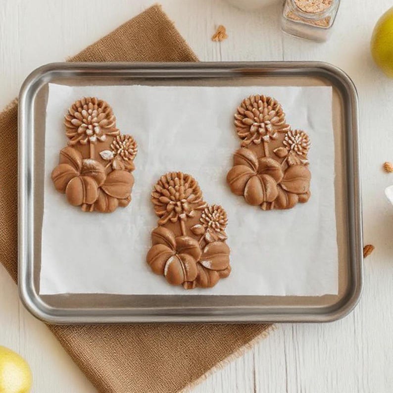 May include: Three brown, flower-shaped cookies on a baking sheet lined with parchment paper. The cookies have detailed floral designs, including petals and smaller flower accents. The baking sheet is silver and sits on a white wooden surface.