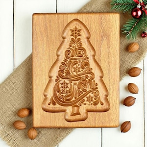 May include: A wooden Christmas tree-shaped mould with detailed floral and swirl carvings. The mould is made of light brown wood and rests on a burlap cloth. Several pecans and a sprig of evergreen with red baubles are also present.